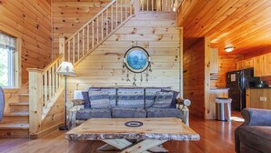 A wooden cabin interior with a staircase, a sofa with patterned cushions, a table with a unique design, and a kitchen area.