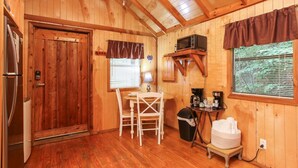 Interior de una cabaña de madera con puerta, una mesa con sillas, microondas, cafetera y ventana con persianas.
