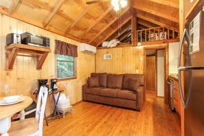 A cozy cabin interior with a wooden ceiling, a sofa, a dining table, and a kitchen area including a refrigerator and a microwave.