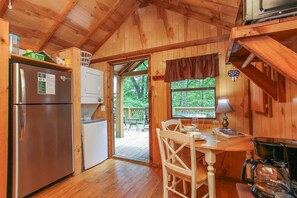 A wooden cabin with a kitchen area, a dining table with chairs, a coffee maker, and a view of the outdoors.