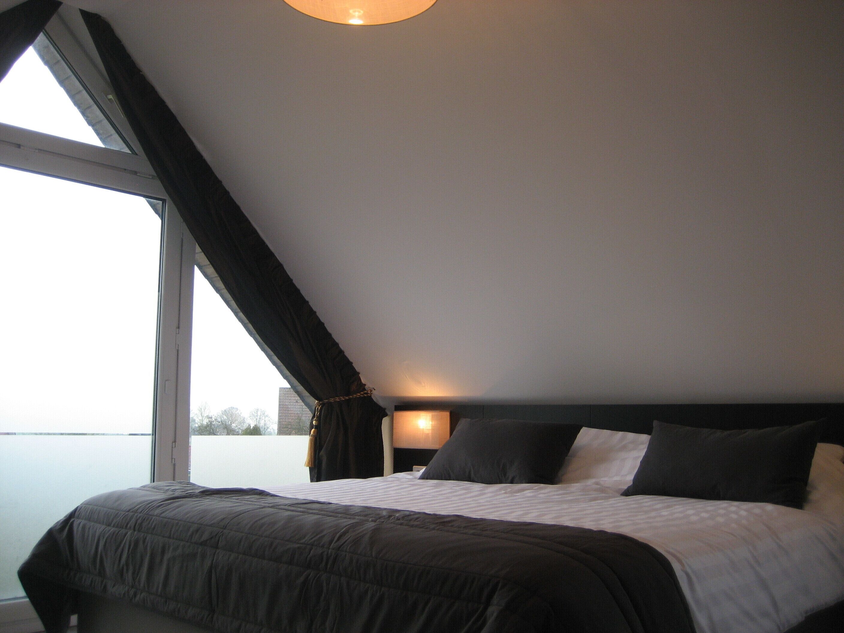 A bedroom with a large window, a bed with a dark headboard, and a bedside lamp.