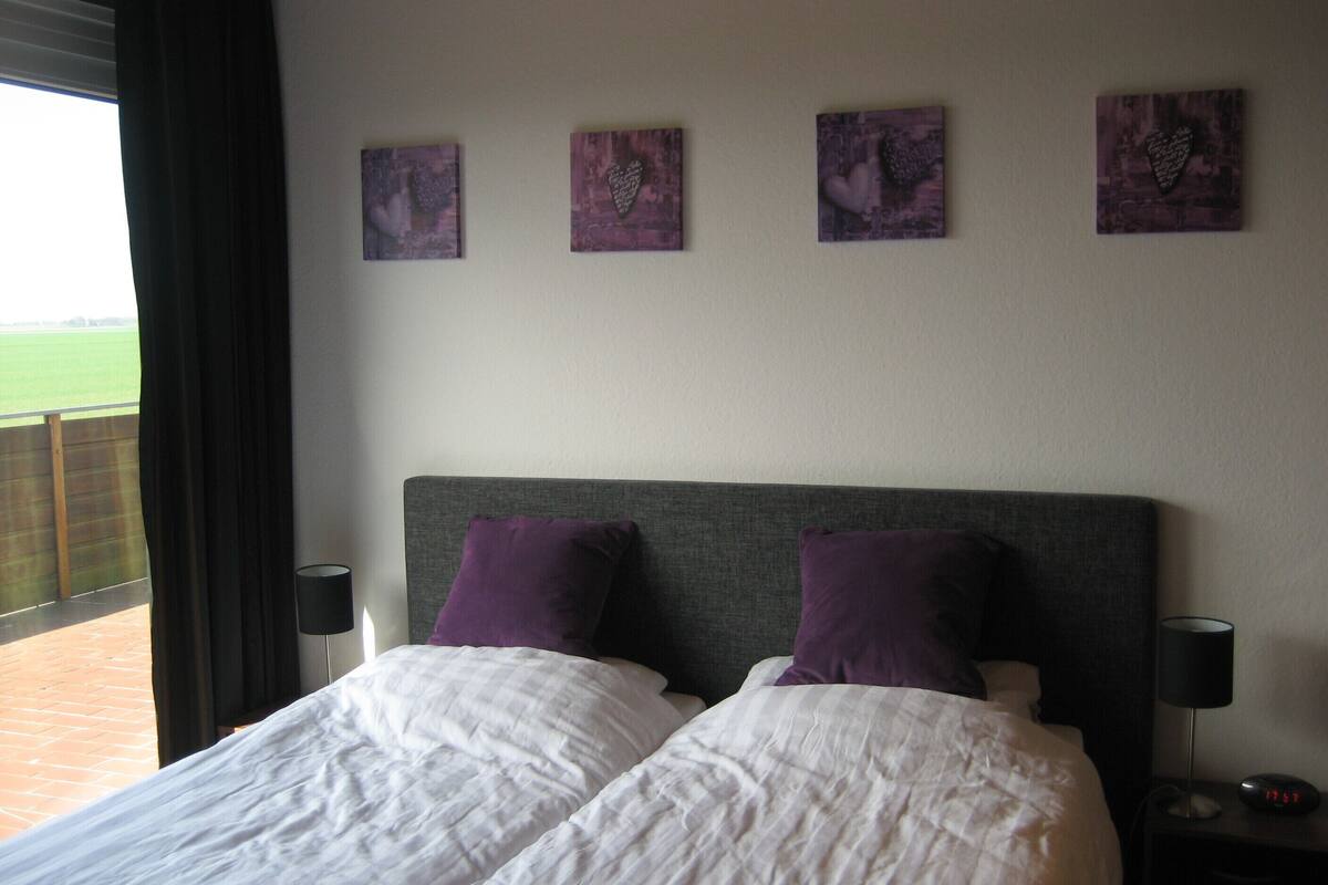 A hotel room with a large bed, two pillows, a nightstand with a lamp, and three wall-mounted artworks.