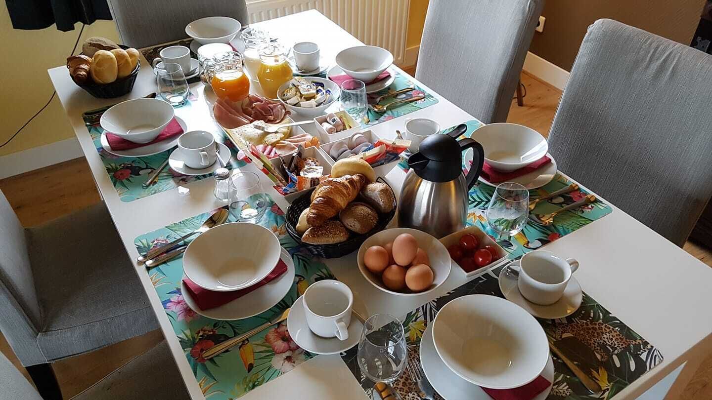 Una mesa de desayuno preparada con varios platos, incluyendo huevos, pan y jugo, en una habitación con sillas grises y un mantel con estampado floral.