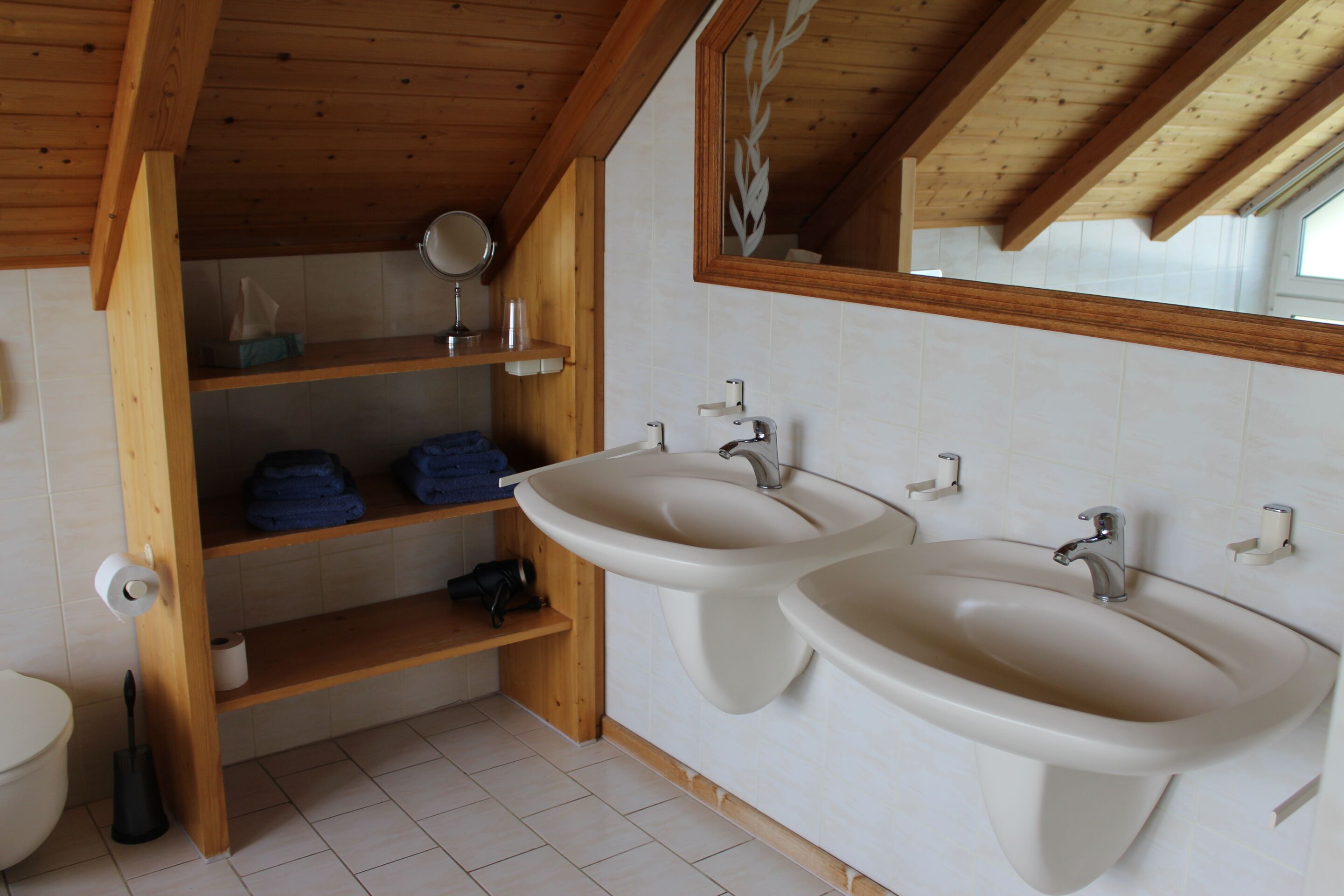 A bathroom with wooden beams, two sinks, a mirror, shelves with towels, and a toilet.