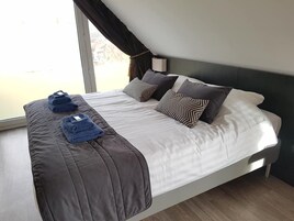 A modern hotel room with a large bed, a window with curtains, and a bedside table with a lamp.