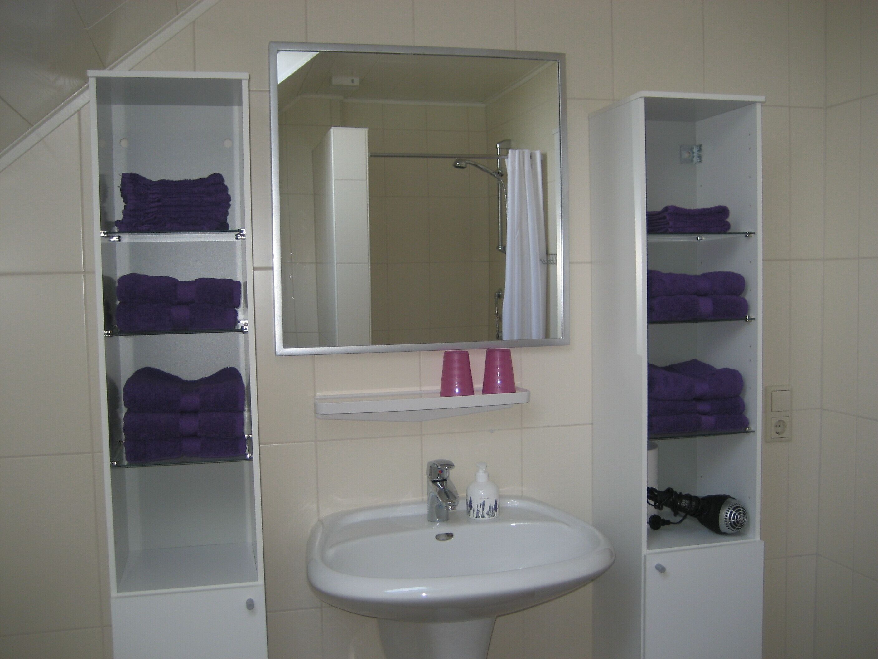 A bathroom with a white sink, a mirror above it, and shelves on either side holding purple towels and pink cups.