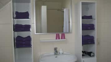 Ein Badezimmer mit einem weißen Waschbecken, einem Spiegel darüber und Regalen an beiden Seiten, auf denen violette Handtücher und rosafarbene Tassen liegen.