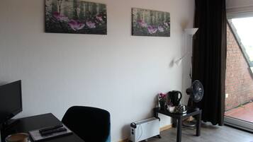 Ein Zimmer mit einem Bett, einem Schreibtisch mit Computer, einem Sessel, einer Heizung, einem Tisch mit Ventilator und zwei Bildern an der Wand.