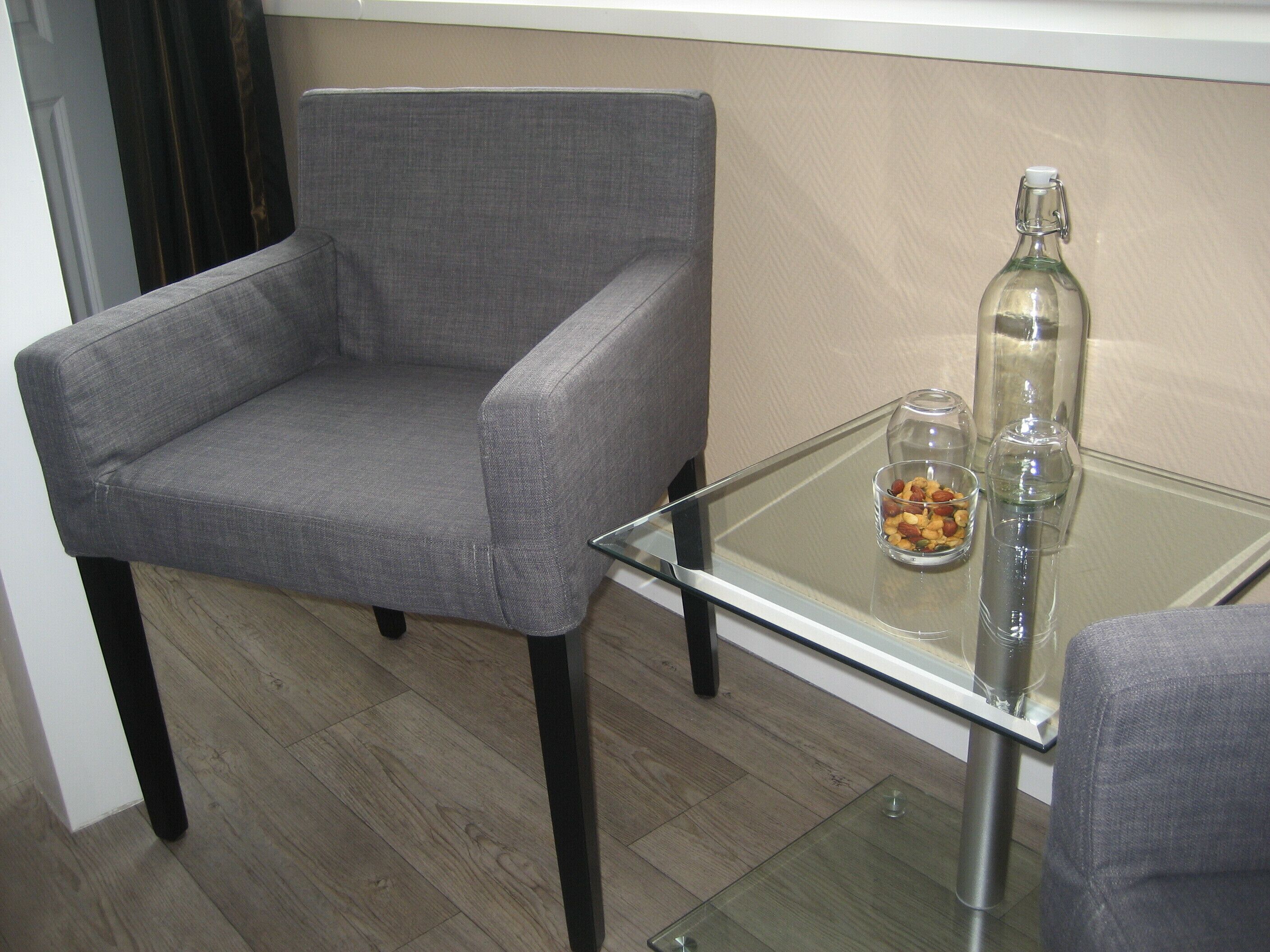 A modern living room with a grey armchair, a glass coffee table with a bottle and glass, and a small bowl of snacks.