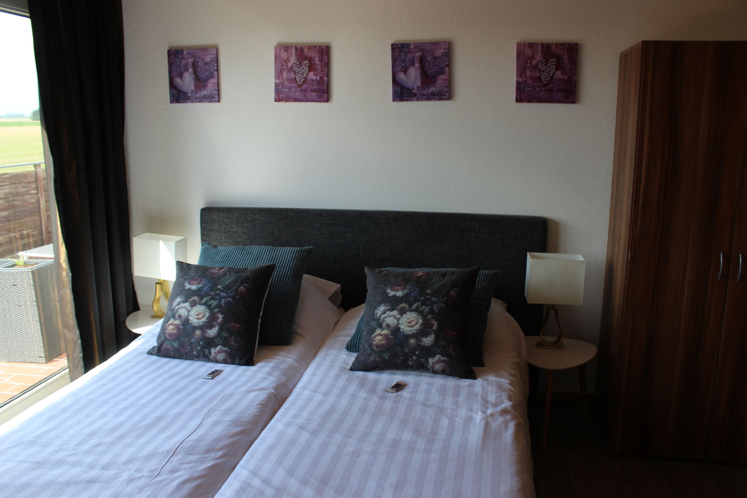 A double bed with floral pillows, a dark headboard, and a view of the outdoors through a window with curtains.