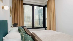 Una habitación de hotel con un ventanal grande, una cama con almohadas verdes y marrones, un ordenador portátil sobre una mesita auxiliar y vistas de edificios altos.