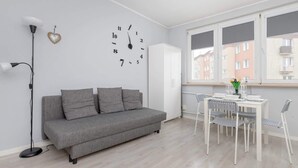 A modern living room with a grey sofa, a white dining table with chairs, a wall clock, and a window with a view of buildings. - Charming Studio with Balcony | Great Location in Gdynia (Gdynia)