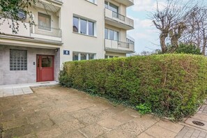 Un immeuble résidentiel à plusieurs étages, avec une porte rouge, un balcon et une haie devant.