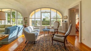 A spacious living room with a high arched ceiling, a large window with a view of greenery, a patterned rug, and a mix of seating including a sofa and armchairs. - Luxury Villa - Unesco Monferrato Langhe Roero with private pool (Asti)