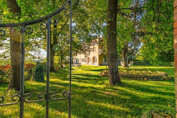 Un lussureggiante giardino con un cancello in ferro battuto, un prato curato e un edificio con uno stile architettonico classico sullo sfondo.