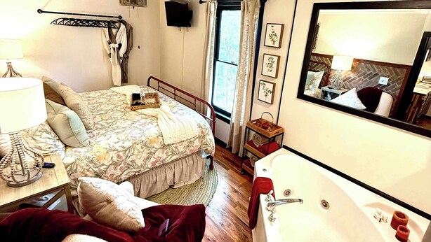 A bedroom with a bed, a bedside table with a lamp, a small table with a decorative item, a wooden floor, a large white bathtub, and a mirror on the wall. - Ridgeway Bed & Brksfst Suite, Full Kitchen, Slps 4 (Eureka Springs)