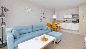 A modern living room with a blue sofa, a white coffee table with fruits, a dining area with yellow chairs, and a kitchen with white cabinets.
