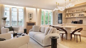 A modern living room with a sofa, coffee table, and dining area. There is a kitchen with a stove and shelves. Large windows provide natural light and a view of greenery outside. - The Lander - Serviced 3BR/3BA - Champs-Elysées (Paris)