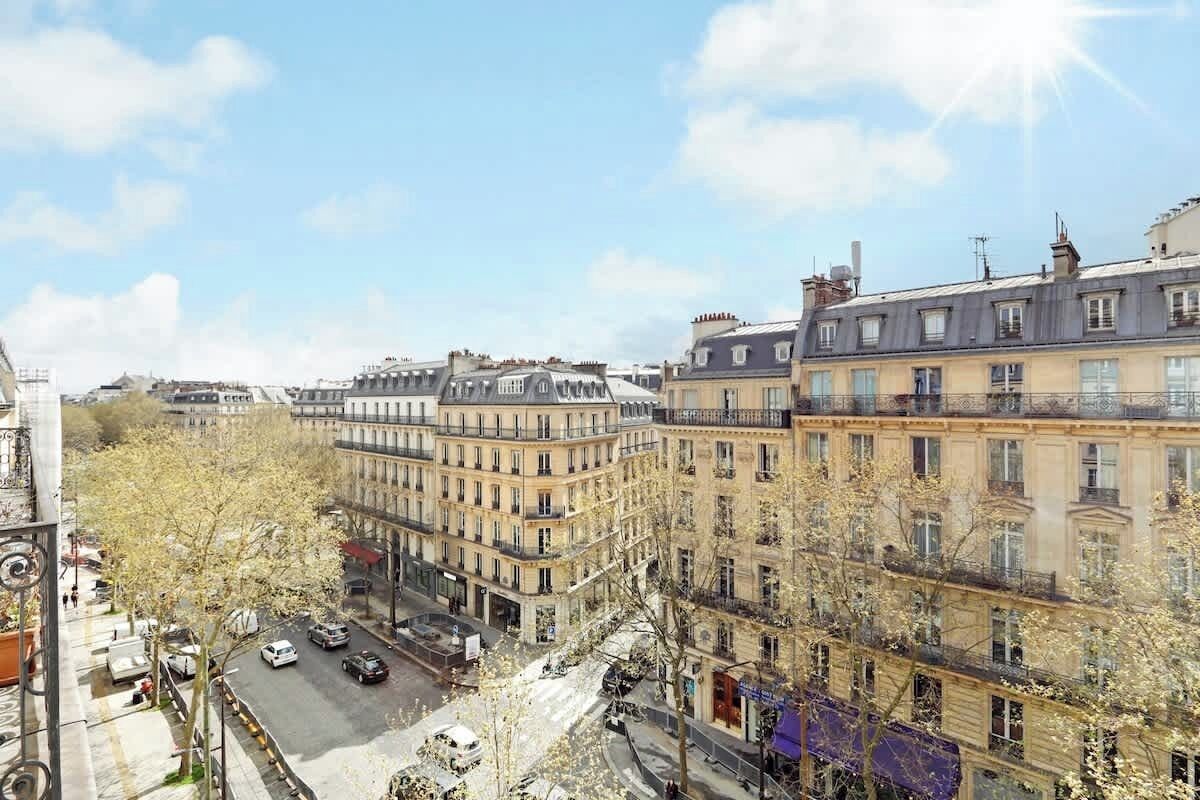 A row of classic European buildings with uniform facades and balconies, a clear sky, and a street with parked cars.