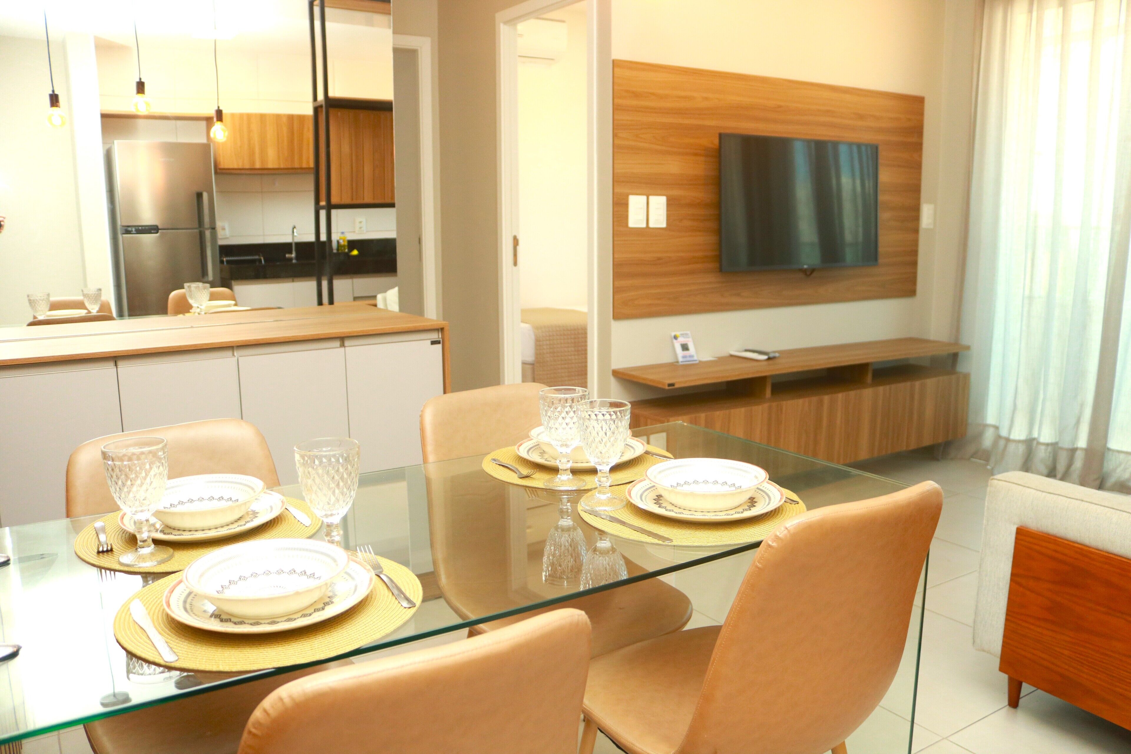 Uma sala de jantar moderna com mesa de vidro, cadeiras bege e televisão montada na parede.