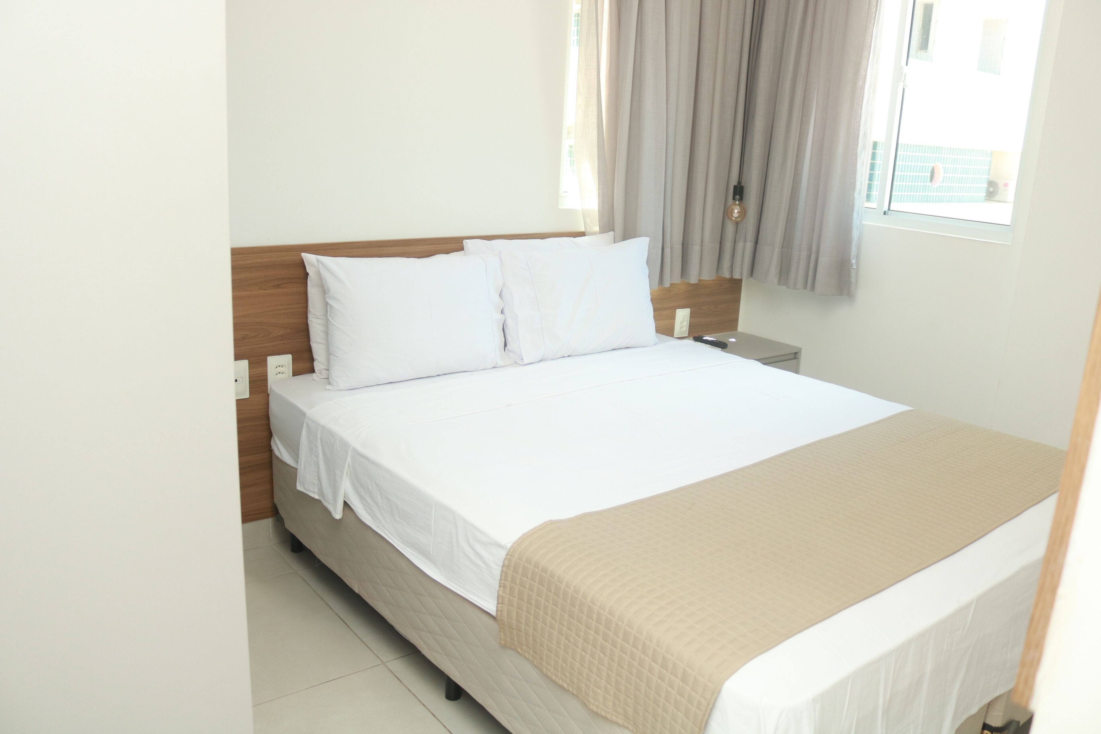 A neatly made bed with white linens and a beige throw blanket, a wooden headboard, a bedside table with a lamp, and a window with curtains.
