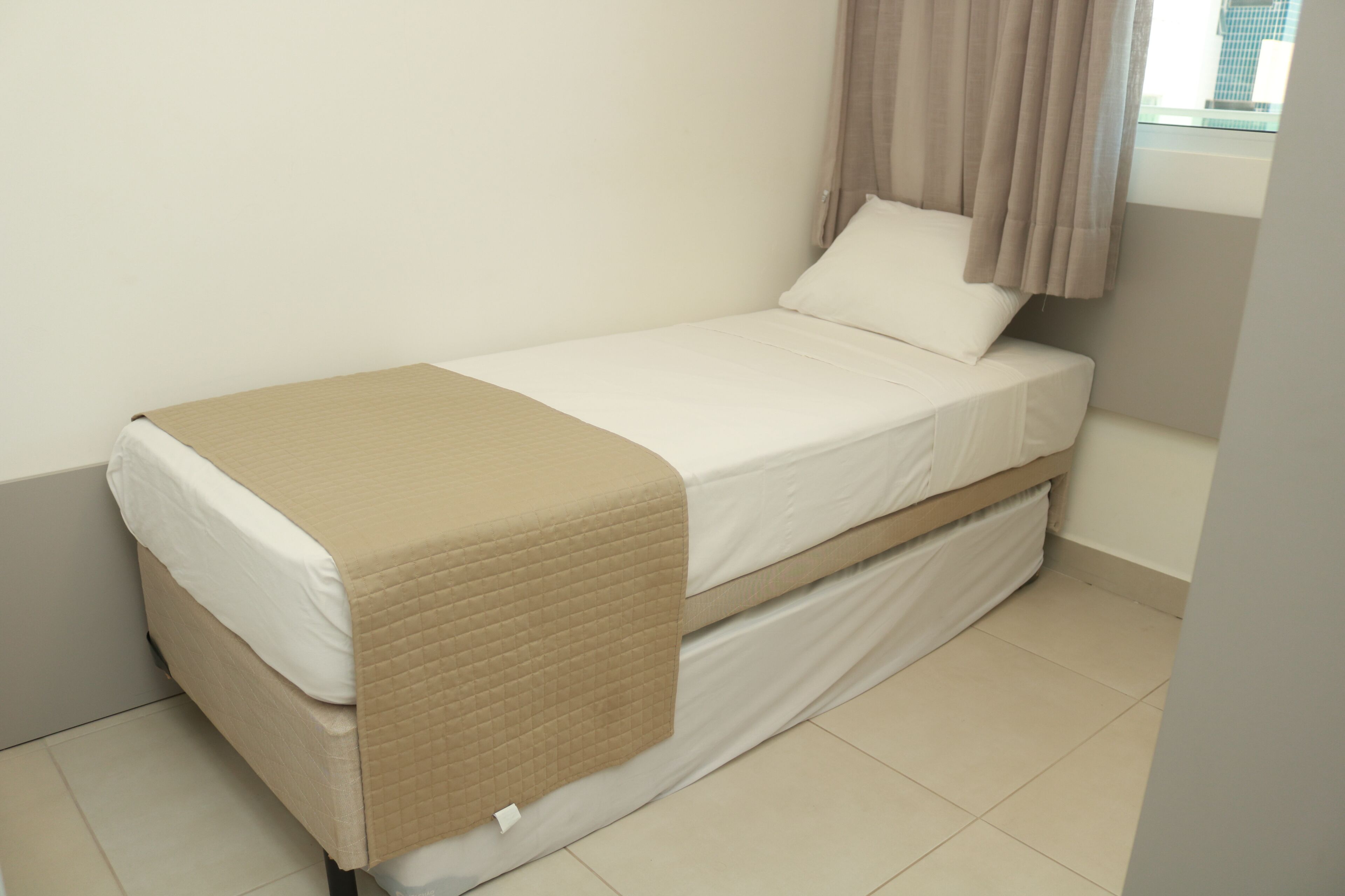 A single bed with a beige quilted cover, white linens, and a window with curtains.