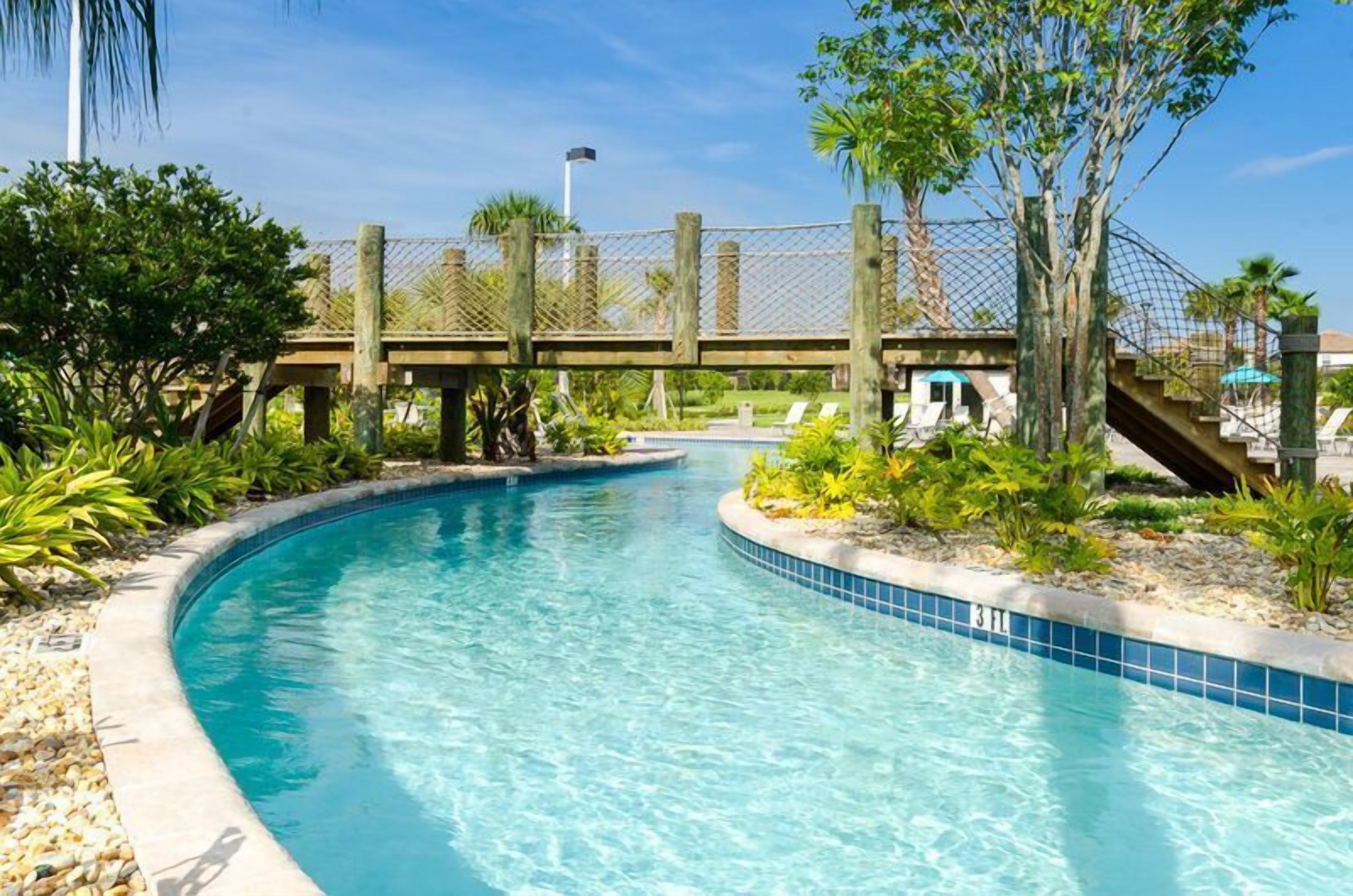 Ein klares Schwimmbecken mit einer Holzbrücke darüber, umgeben von tropischen Pflanzen und blauem Himmel.