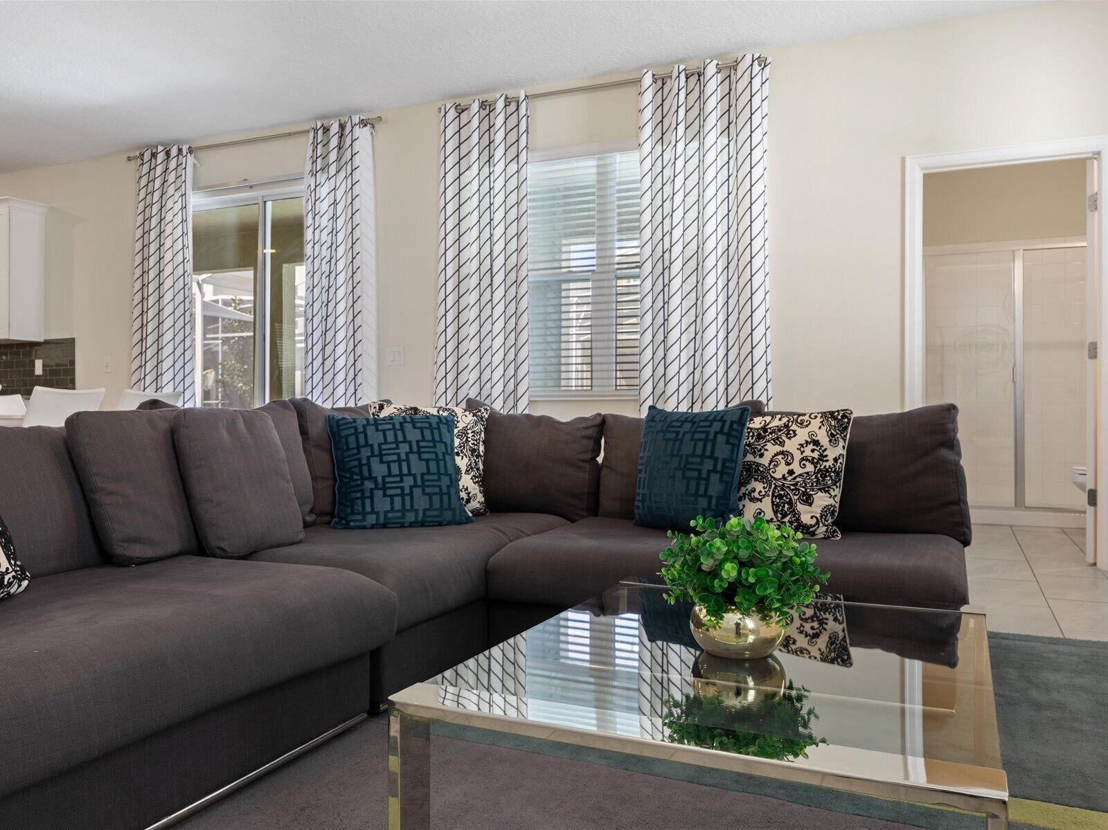 A modern living room with a sectional sofa, patterned cushions, a glass coffee table, and a view of a bathroom through a window with blinds.