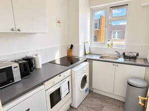 A compact kitchen with a microwave, toaster, and washing machine, a window with a view of buildings, and a sink with a faucet. - The Elden - No.9 (London)