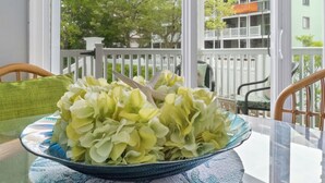 Une table sur laquelle se trouve un vase en verre rempli de fleurs, une chaise et une vue sur un balcon avec une balustrade et de la verdure.