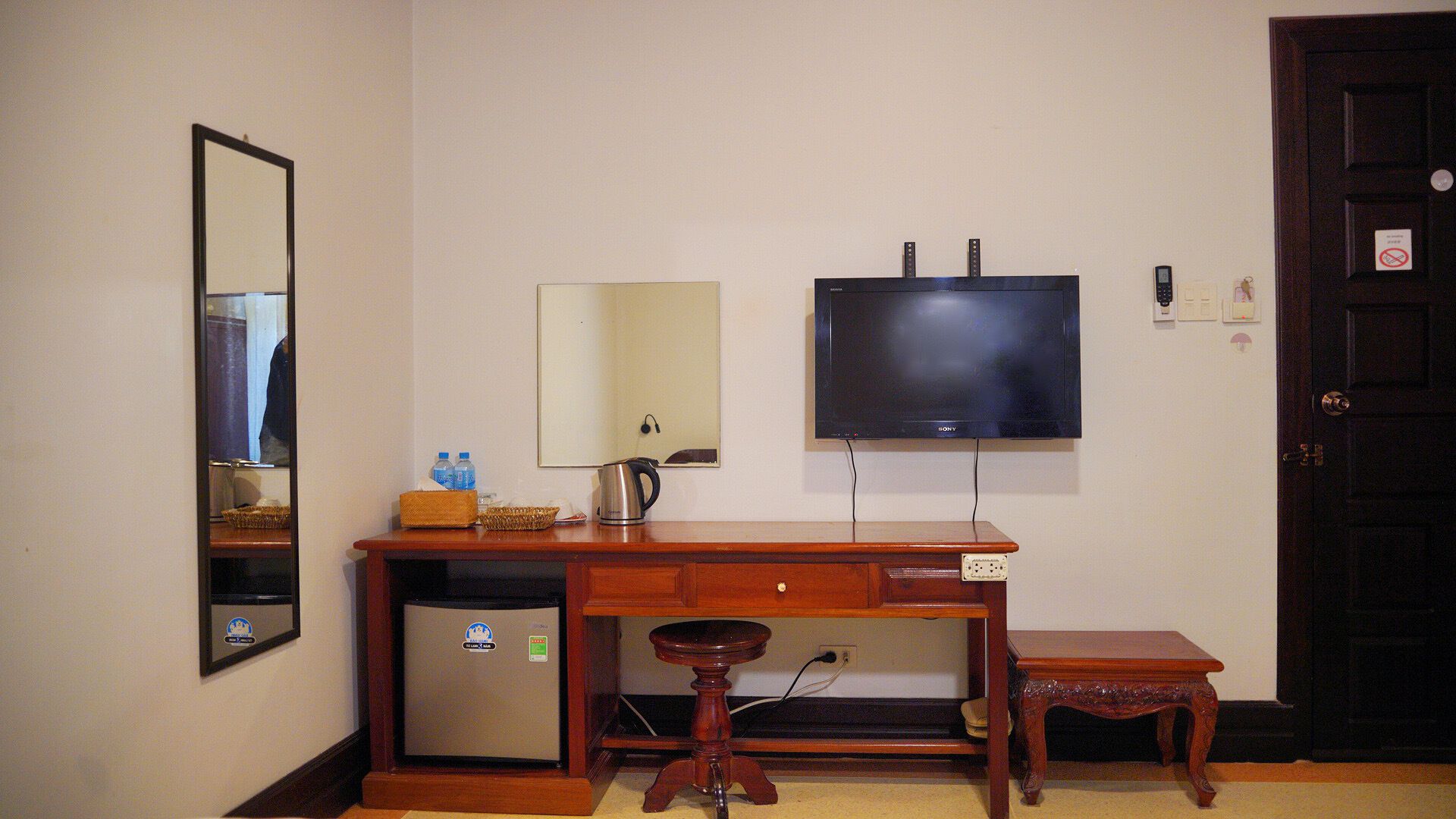 A compact room with a wooden desk, a small stool, a wall-mounted TV, a mirror, and a door.