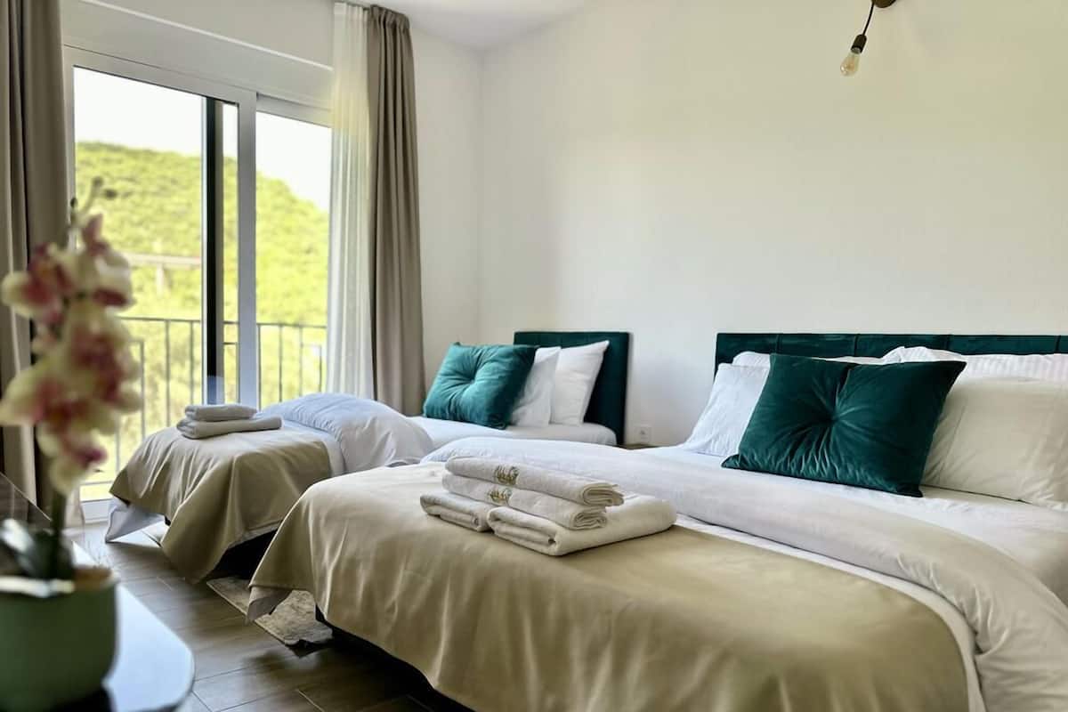 A hotel room with two beds, each with white linens and green pillows, a large window with curtains, and a potted plant on the left side.