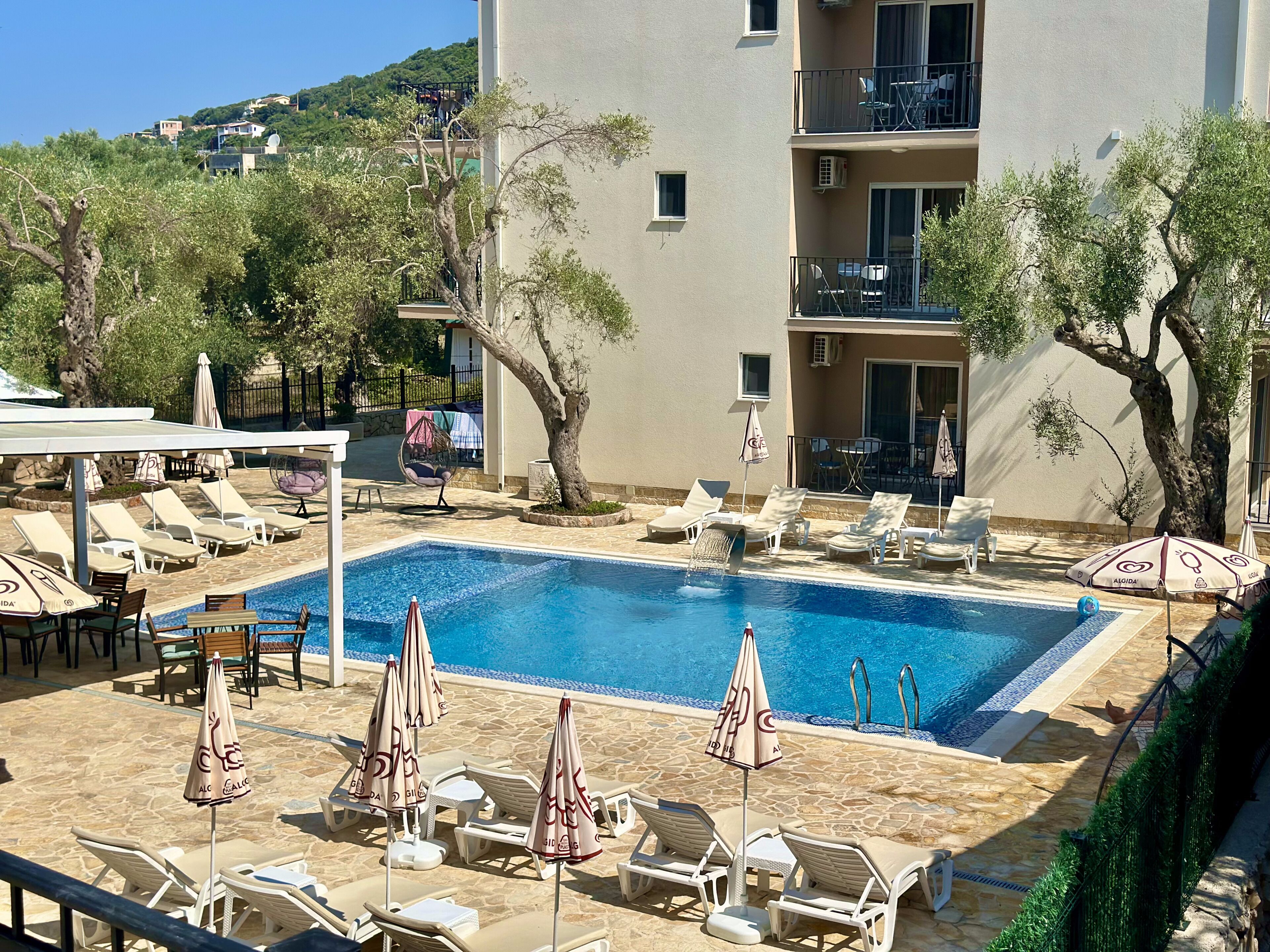 Piscina con sillas de descanso y sombrillas, rodeada de árboles y un edificio con balcones.