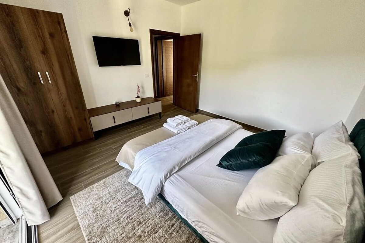 A modern bedroom with a large bed, a wooden wardrobe, a flat-screen TV, and a small bedside table.