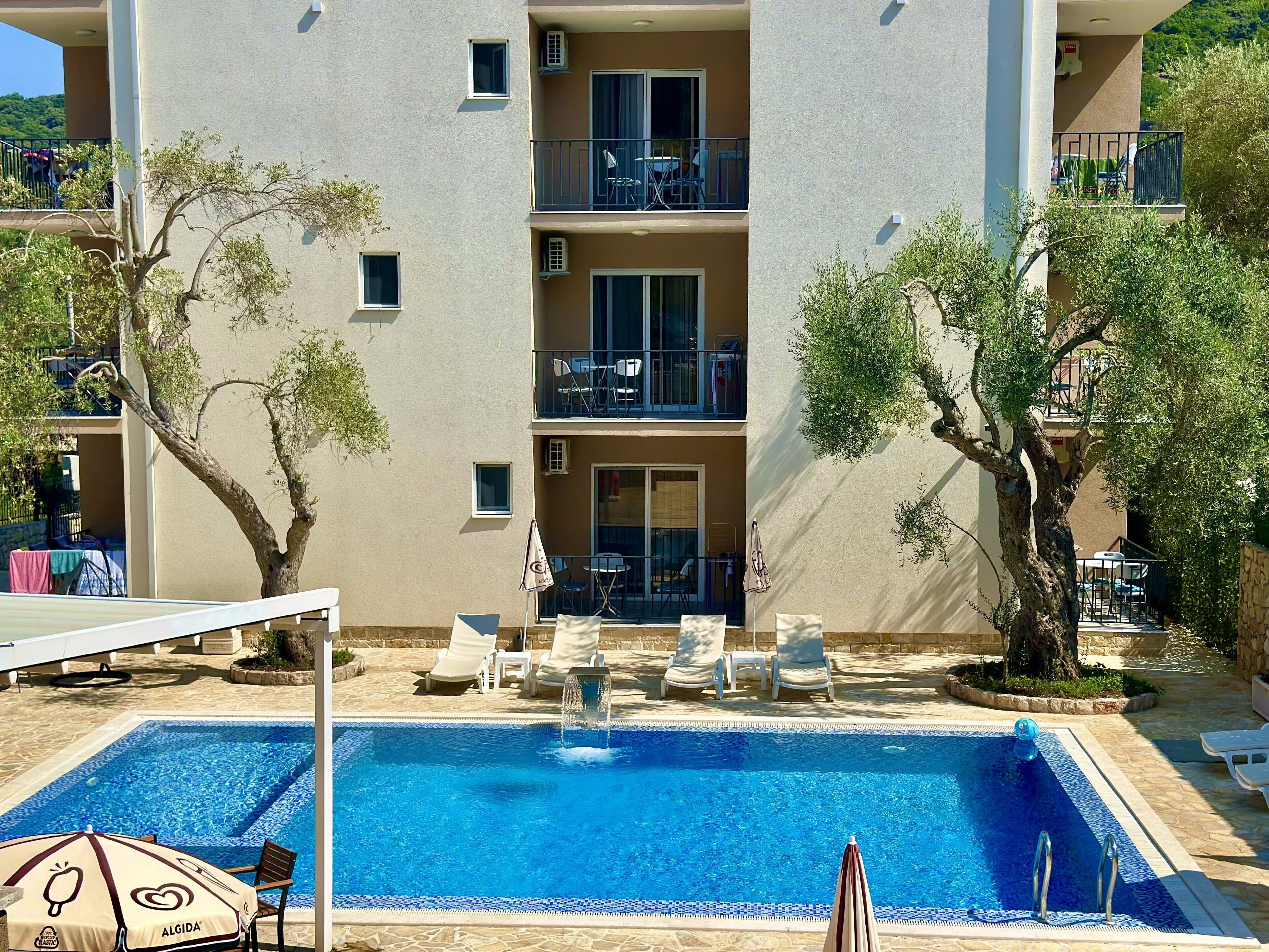 A pool area with lounge chairs and umbrellas, surrounded by trees and a building with balconies.