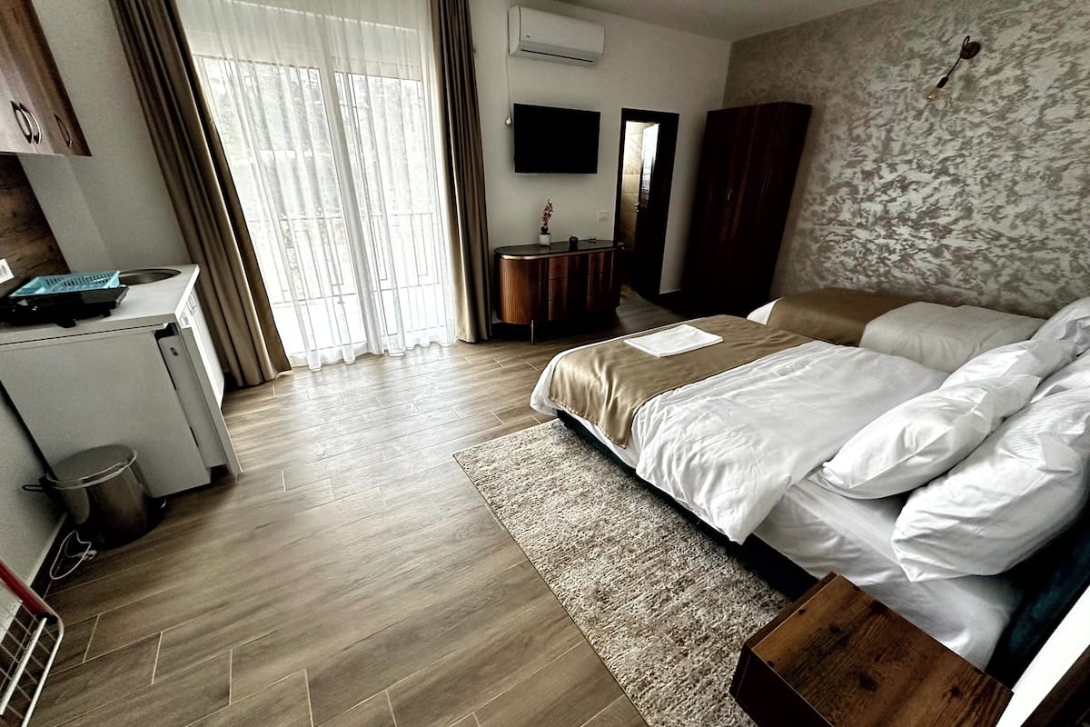 A hotel room with a bed, a desk, a chair, a small trash bin, and a window with curtains.