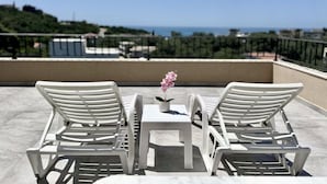 A terrace with two white lounge chairs and a small table, overlooking a scenic view of the sea and greenery. - Akhdar Apartments (Utjeha)