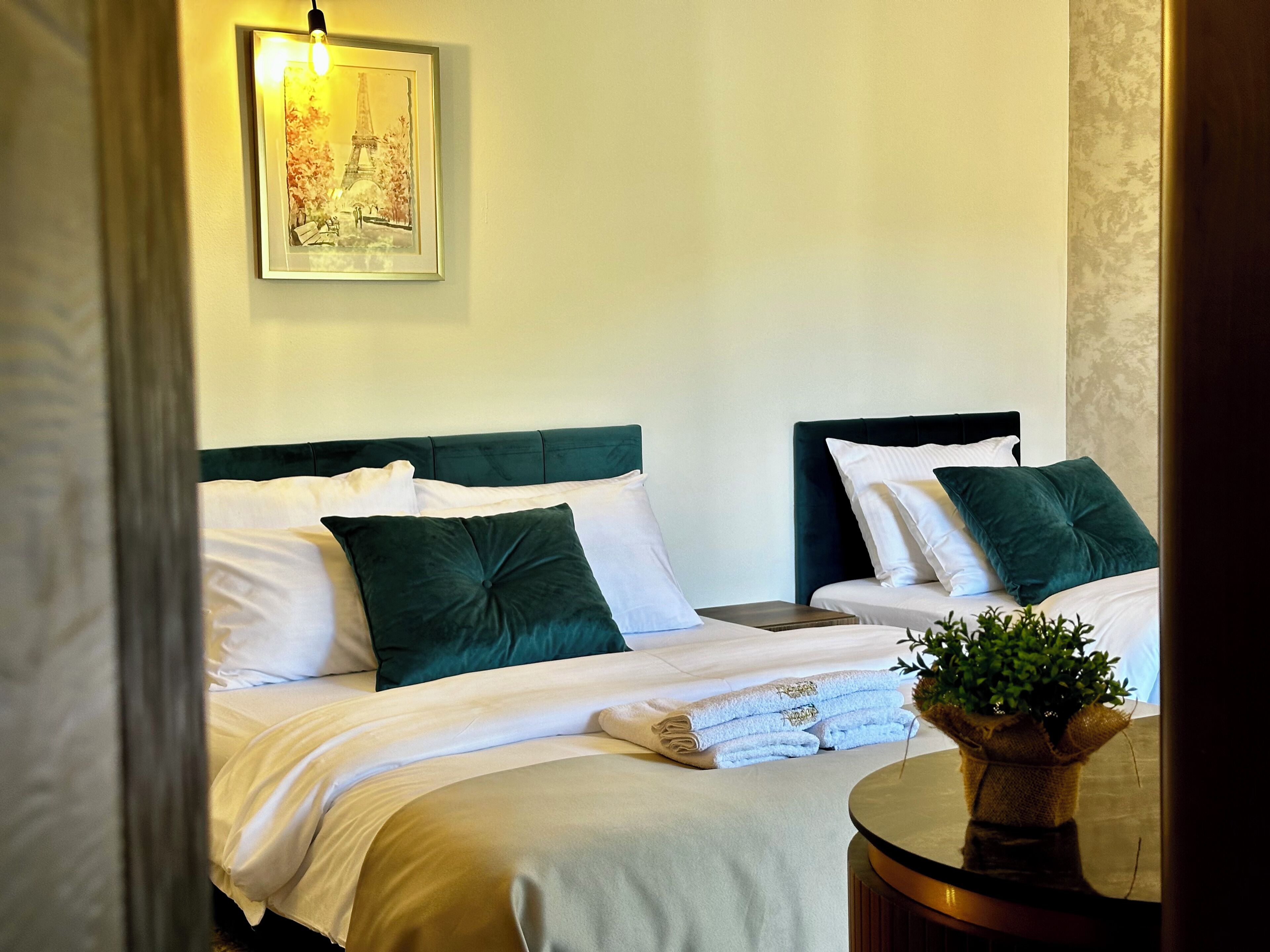A hotel room with two beds, each with green headboards and white bedding. There is a framed picture of the Eiffel Tower on the wall above the beds.