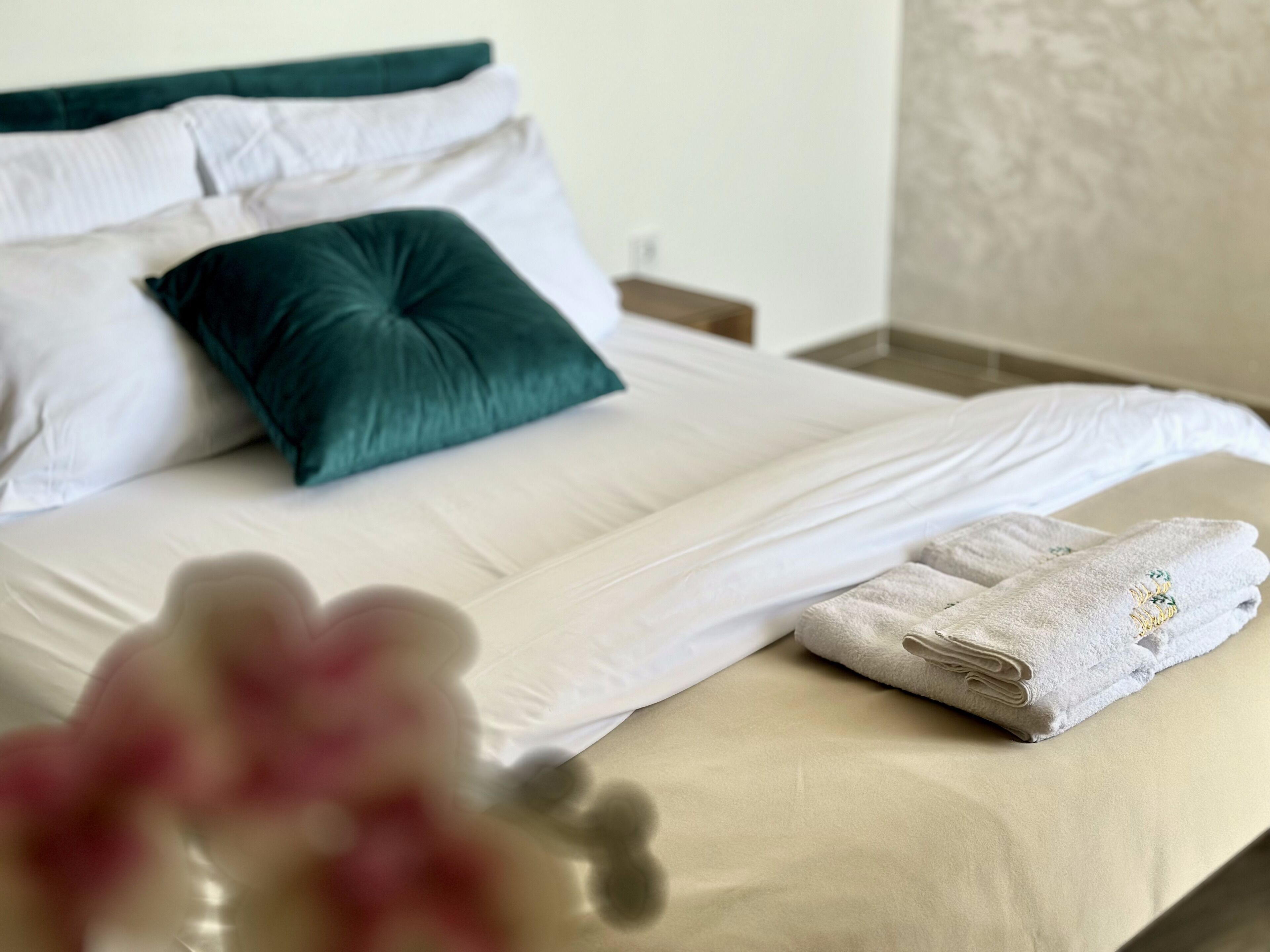 Een net opgemaakt bed met witte lakens en een turquoise kussen, een opgevouwen handdoek met een bladpatroon, en een vaas met roze bloemen op de voorgrond.