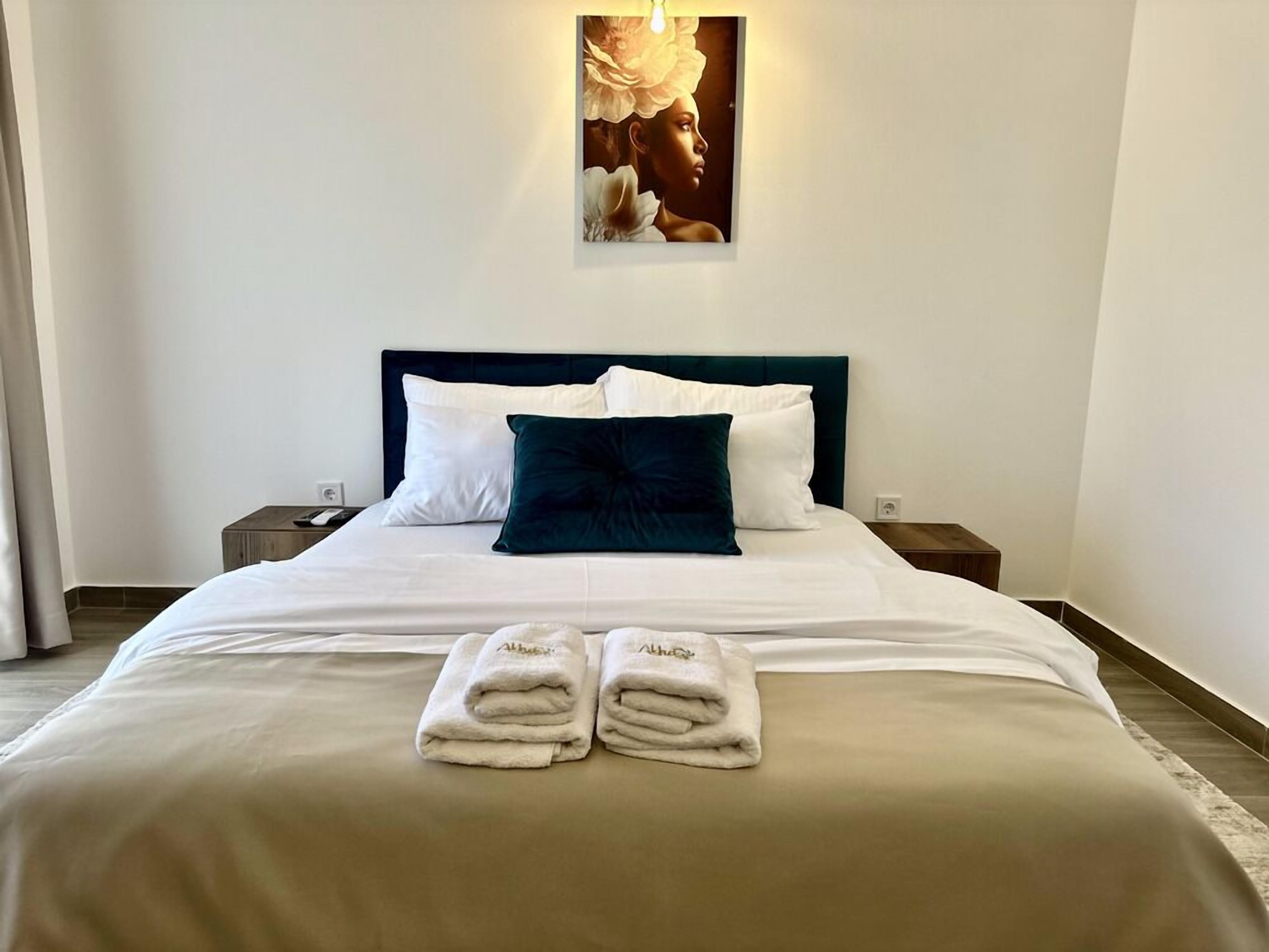 A neatly made bed with white and dark blue pillows, two folded towels, and a wall-mounted light fixture above.