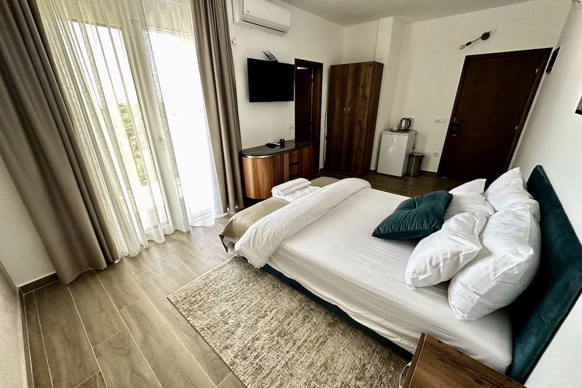 A hotel room with a large bed, a flat-screen TV, a wardrobe, a small kitchenette, and a window with curtains.
