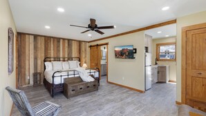 Un dormitorio con cama, cabecera de madera, ventilador de techo y vista a la cocina.