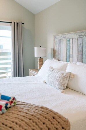 A bed with a white comforter and two pillows, a lamp on a bedside table, and a view of the outdoors through a window with curtains. - Wave After Wave | Dogs OK, Walk to Beach, Pool (Corolla)