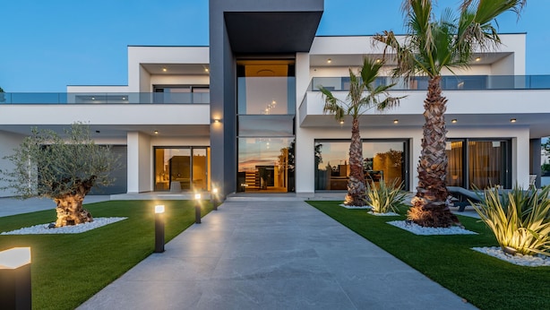 A modern two-story house with a well-lit entrance, a paved driveway, and landscaped with palm trees and shrubs.