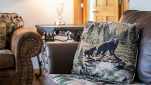 A living room with a brown leather sofa, a patterned throw pillow, a table with a lamp, and a wooden door in the background. - New! Private 2BR Cabin w/Hot Tub*Central Location (Sevierville)