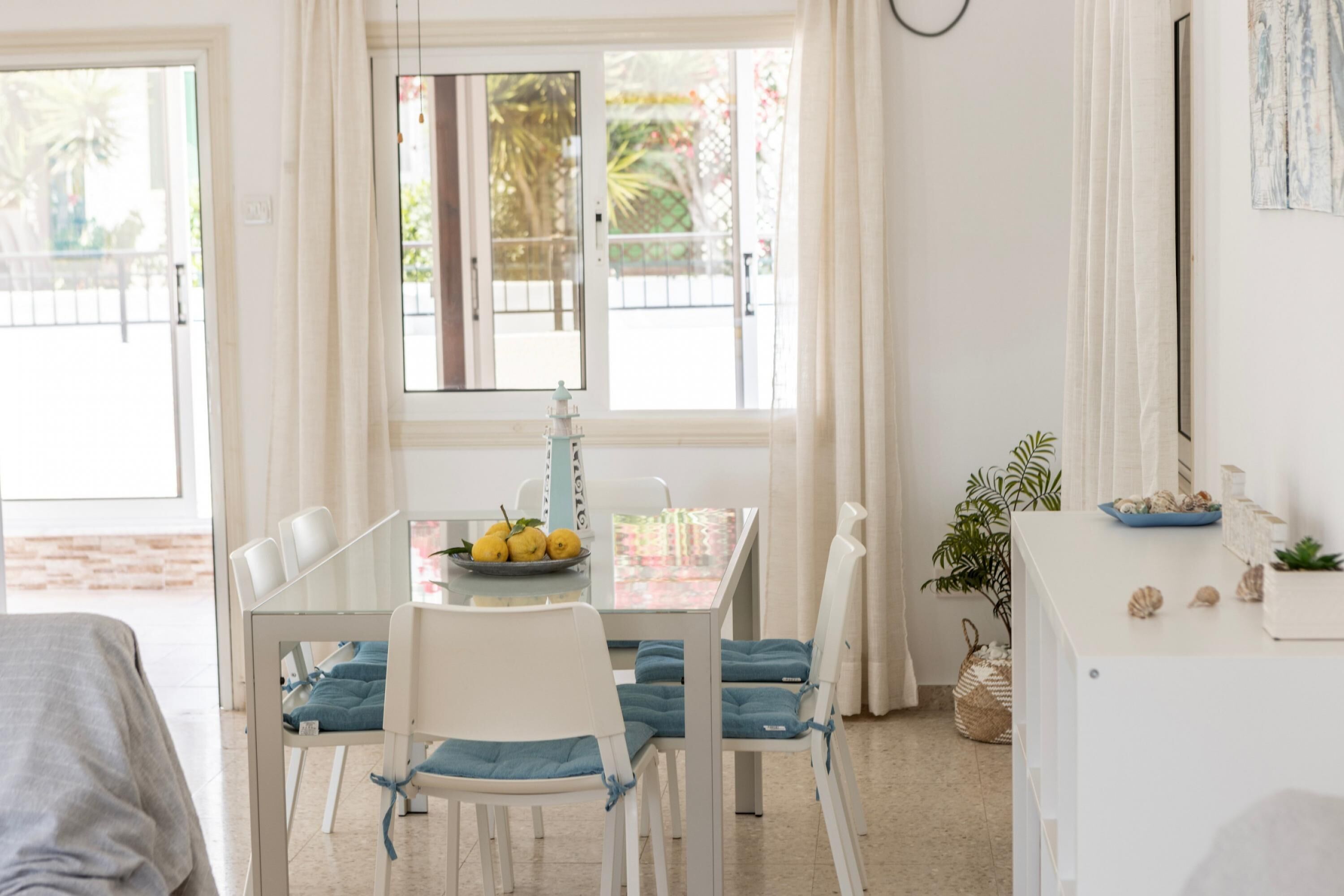 A bright dining area with a table set for two, chairs with blue cushions, a window with a view of greenery, and a small plant on the side.