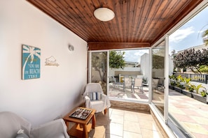 A covered patio area with a wooden ceiling, a white chair, a small table, and a glass door leading outside. - Ayia Thekla Sea Breeze Villa (Sotira)