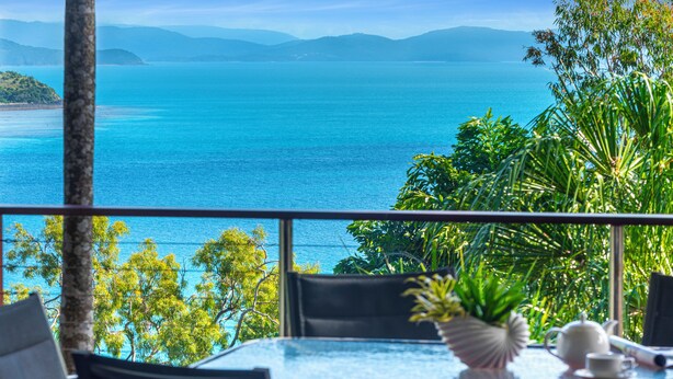 A table with chairs and a potted plant, overlooking a turquoise sea and distant mountains.