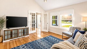 Una sala de estar con televisor de pantalla plana, un sofá, un tapete estampado y vistas a un paisaje verde a través de amplios ventanales.