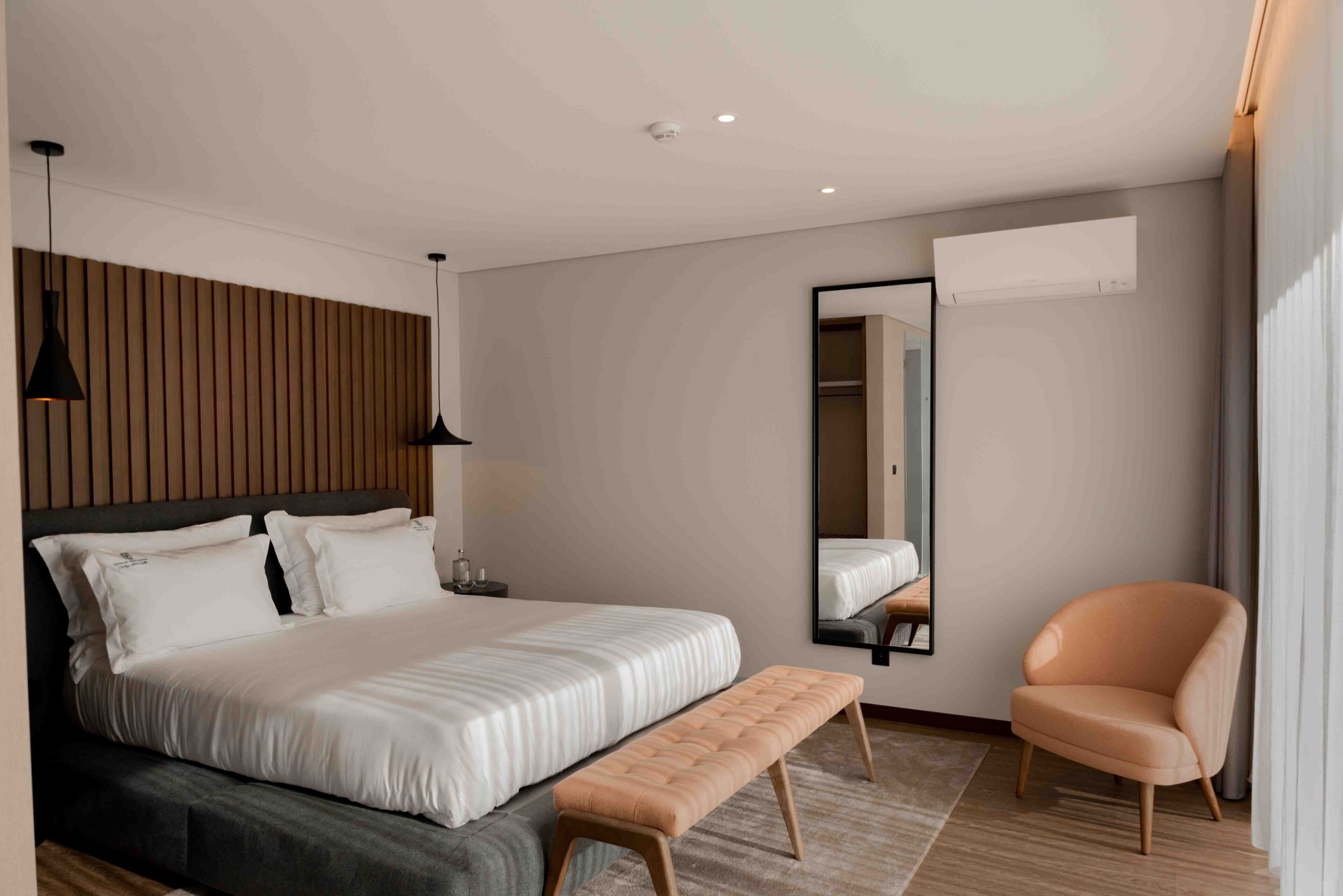 A modern bedroom with a large bed, a bench, a chair, a mirror, and a wall-mounted air conditioner.